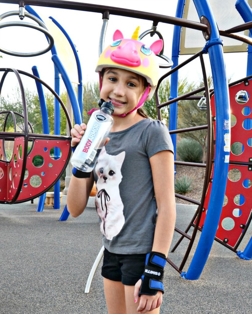 Outdoor exercise with essentials for kids at the park include protective gear, differing forms of transportation and drinks for hydration.